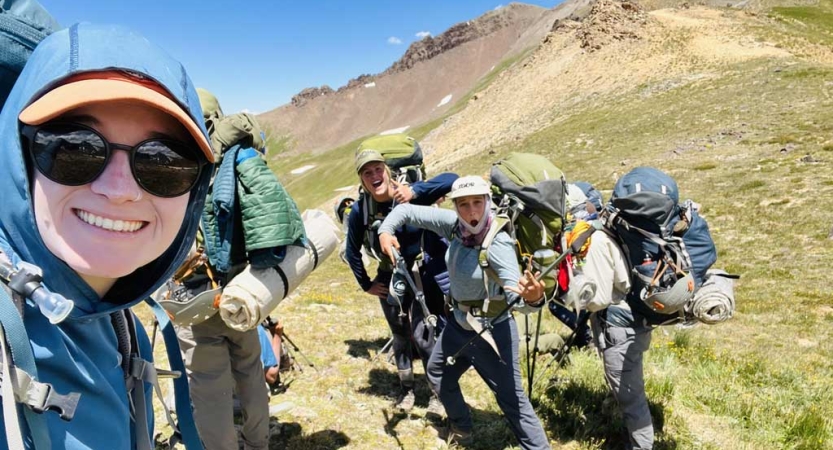 a group of people wearing hiking gear stand in a green alpine meadow. Some of them make silly poses. 